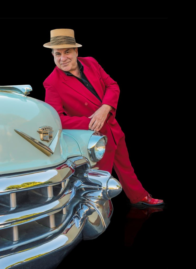 photo of blues piano player Mitch Woods leaning on the bonnet of a car and wearing a bright red suit